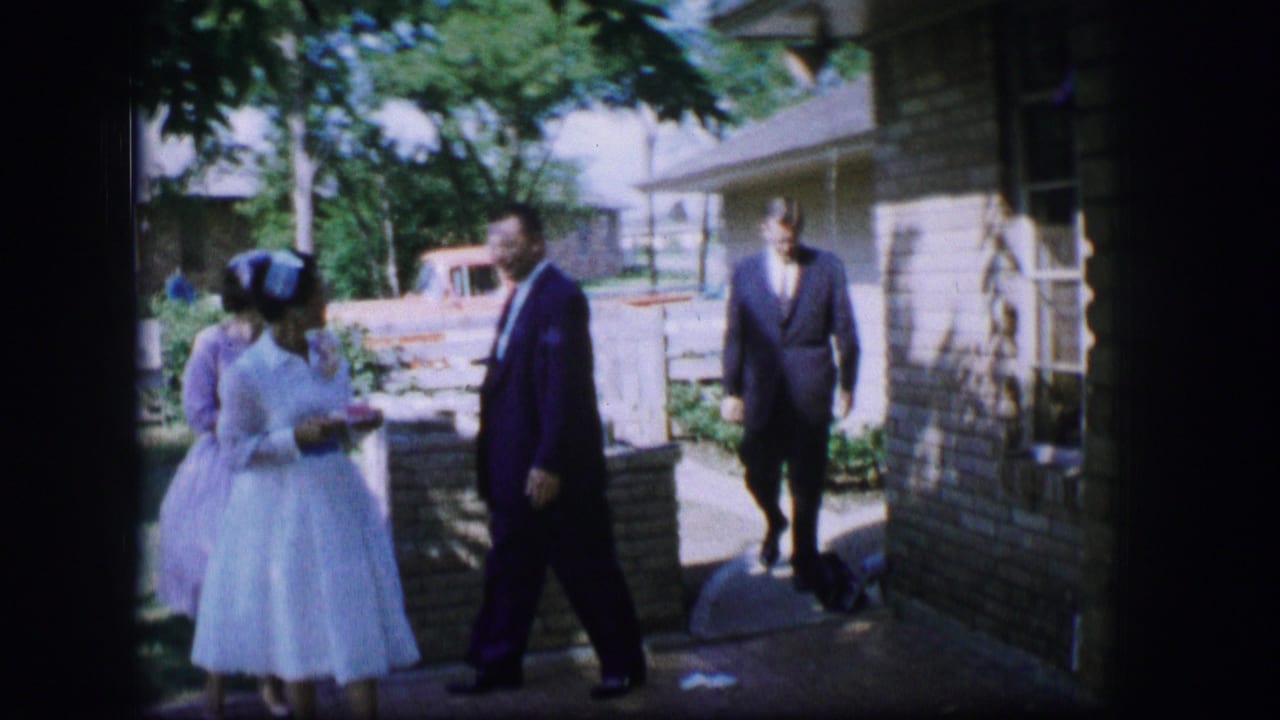 In 1962 Moberly, Missouri, a bride in a white gown stands outside while suited men move through a shaded yard near a house and stone wall.