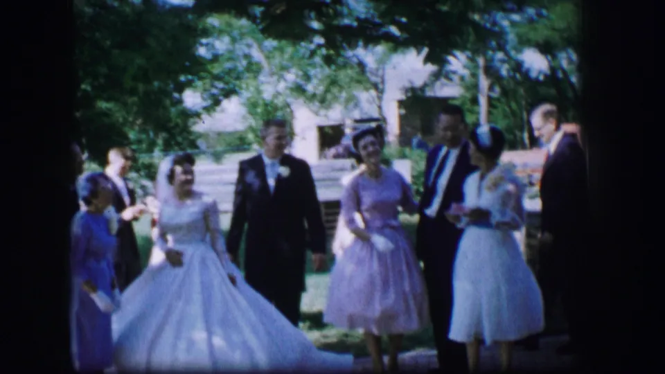 In the Missouri wedding scene, the bride smiles beside a suited man as women in pastel dresses and other guests stand in the shaded yard.