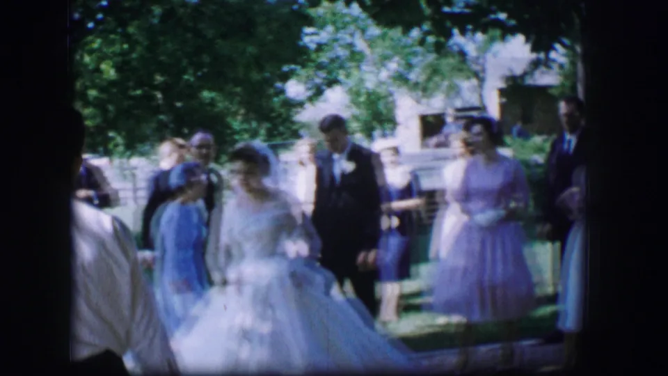 The bride and wedding guests stand on a lawn in Moberly, Missouri, with pastel dresses and dark suits visible in the 1962 home movie frame.