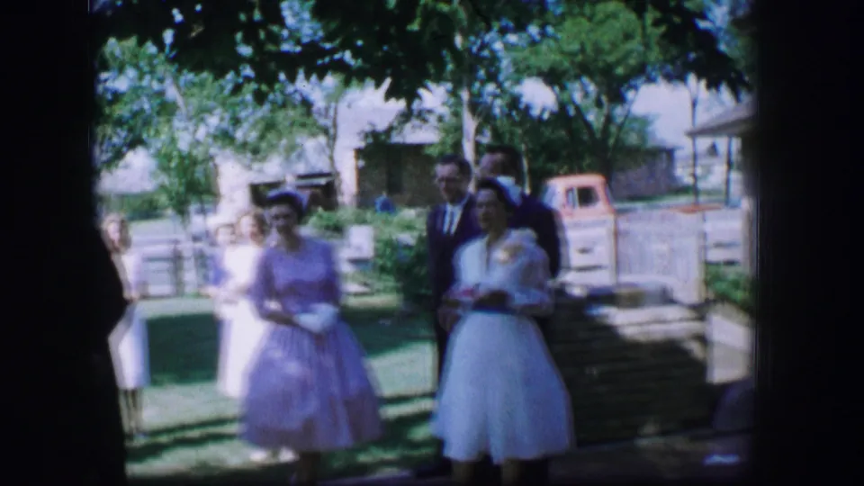 Women in pastel formal dresses stand with suited guests during the 1962 Moberly wedding gathering, with a fence and parked vehicle in the background.