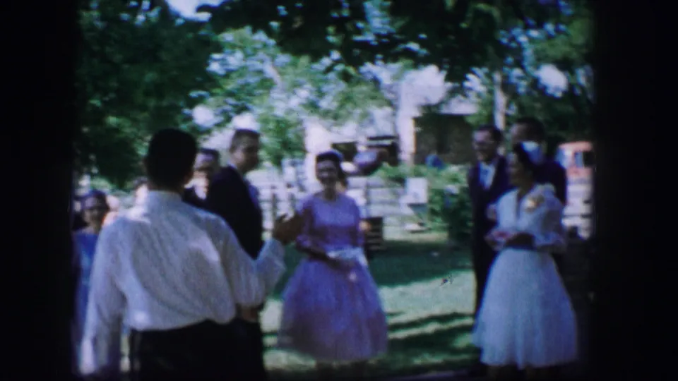 A man in a white shirt stands with his back to the camera while wedding guests and attendants gather on a Moberly lawn in 1962.