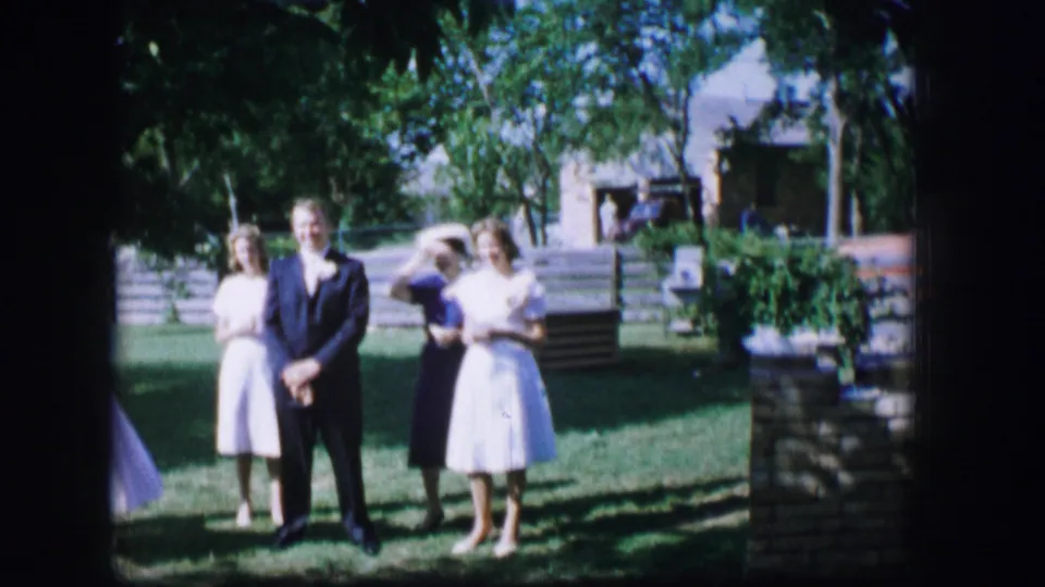 1962 Moberly, Missouri wedding frame showing a suited man and women in dresses standing in a shaded yard.