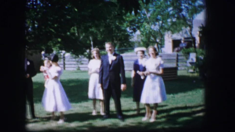 1962 Moberly, Missouri 8mm frame with guests in pastel dresses and a dark suit gathered on a tree-shaded lawn.