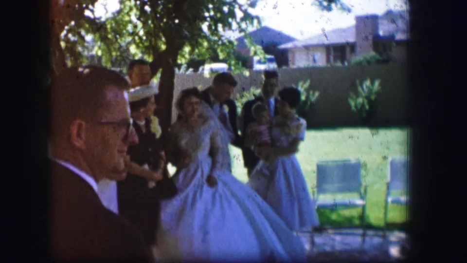 A home-movie view of a 1962 Moberly wedding shows the bride, groom, and guests in formal clothing on a sunlit lawn.