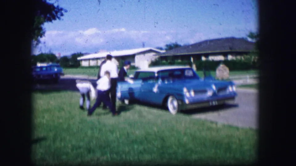 A blue station wagon is parked outside a suburban home as formally dressed people gather nearby in Moberly, Missouri, 1962.