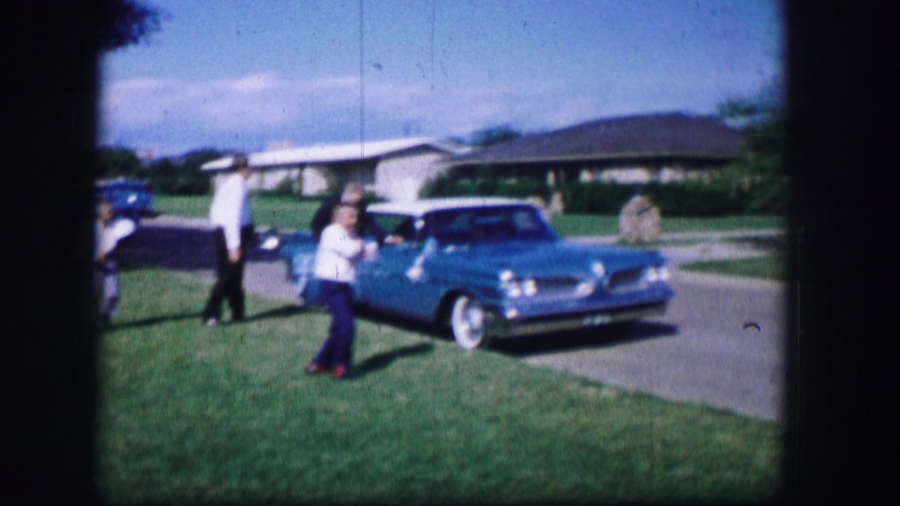 In 1962 Moberly, Missouri, children play on a front lawn beside a bright blue car while adults stand near a driveway and single-story homes.