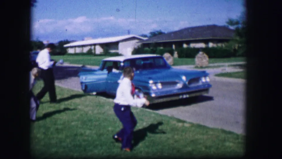A child moves across a Moberly lawn near adults and a blue early-1960s car parked by a driveway, with houses in the background.