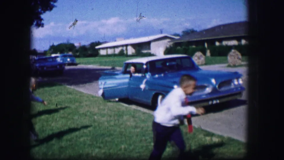 A child moves low across the foreground while a bright blue car with an open door is parked near a suburban lawn in Moberly, Missouri.