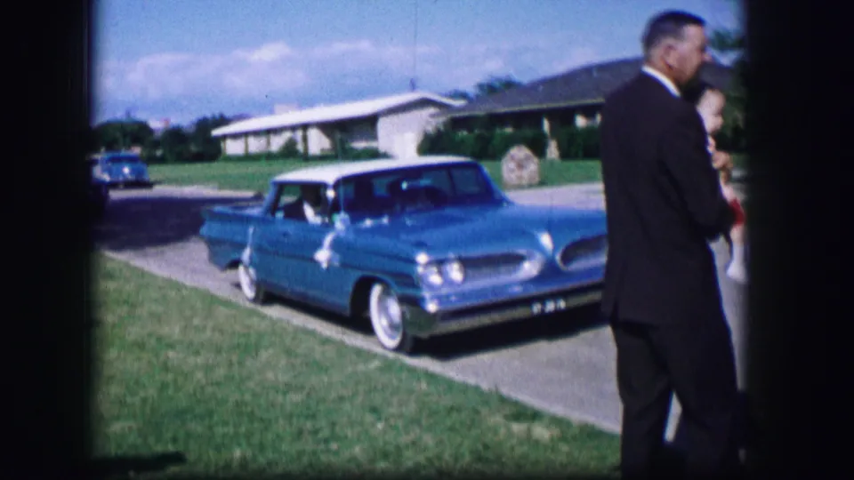 Adults in dress clothing stand at the edge of the frame as a blue vintage car sits in a driveway near a lawn and houses in 1962 Moberly.