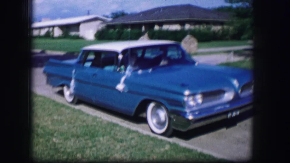 The blue Pontiac moves forward along the curb in 1962 Moberly, Missouri, with lawns and low suburban houses behind it.