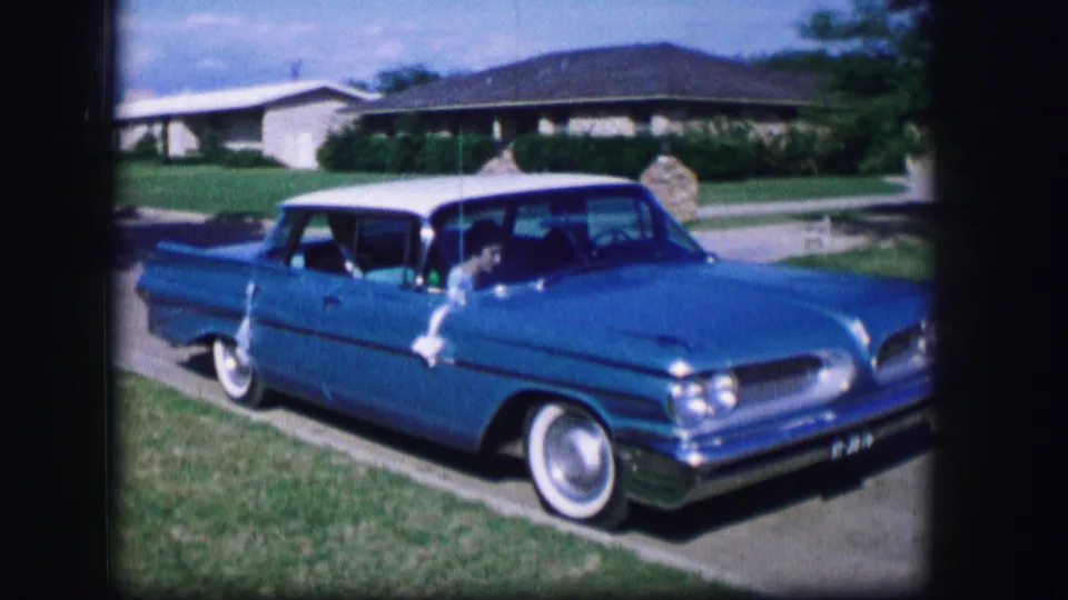 A close view shows the blue Pontiac passing by with chrome trim, whitewall tires, and suburban houses in the background.