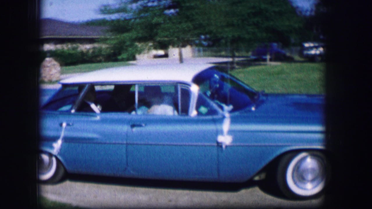 A blue classic car drives past ranch houses in the supplied 1962 Moberly, Missouri home-movie setting.