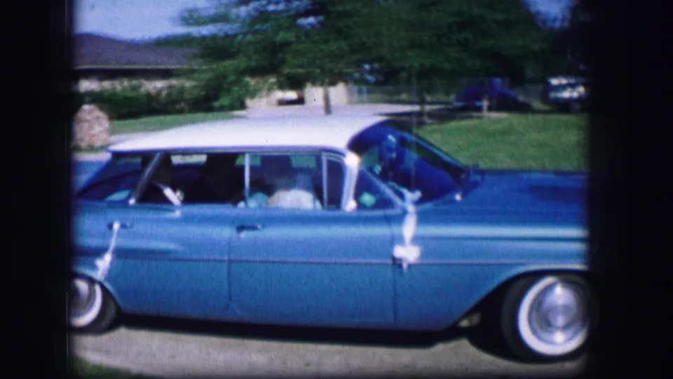 A blue classic car drives past ranch houses in the supplied 1962 Moberly, Missouri home-movie setting.
