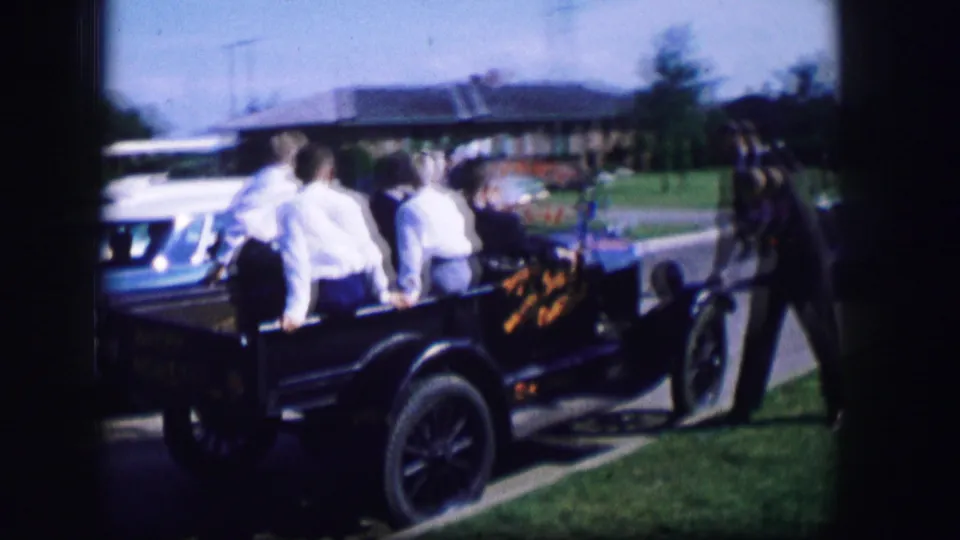 Several young adults sit in a white open-top vehicle as a blue car is nearby on a suburban street.