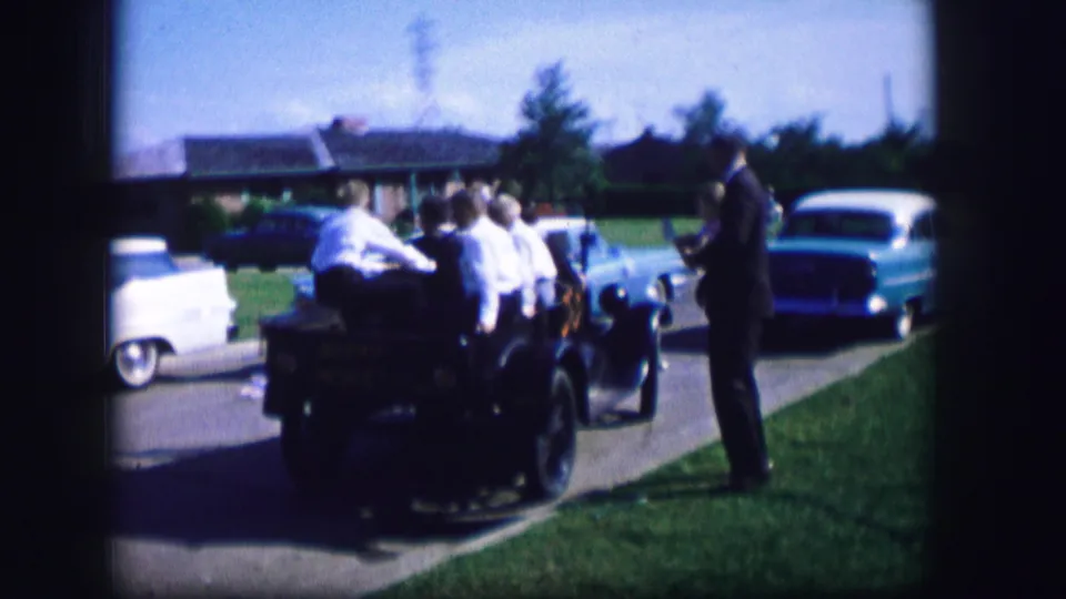 The open-top car carrying young adults is stopped or moving slowly near a suited man and the blue car in the street.