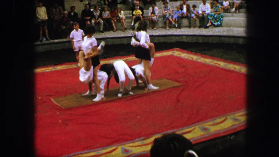 Young performers continue an outdoor gymnastics routine on mats in Austria, with seated spectators visible behind the red carpeted stage.