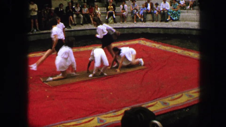 A mid-routine home-movie frame shows amateur gymnasts on mats, with one performer moving across the formation as spectators look on.
