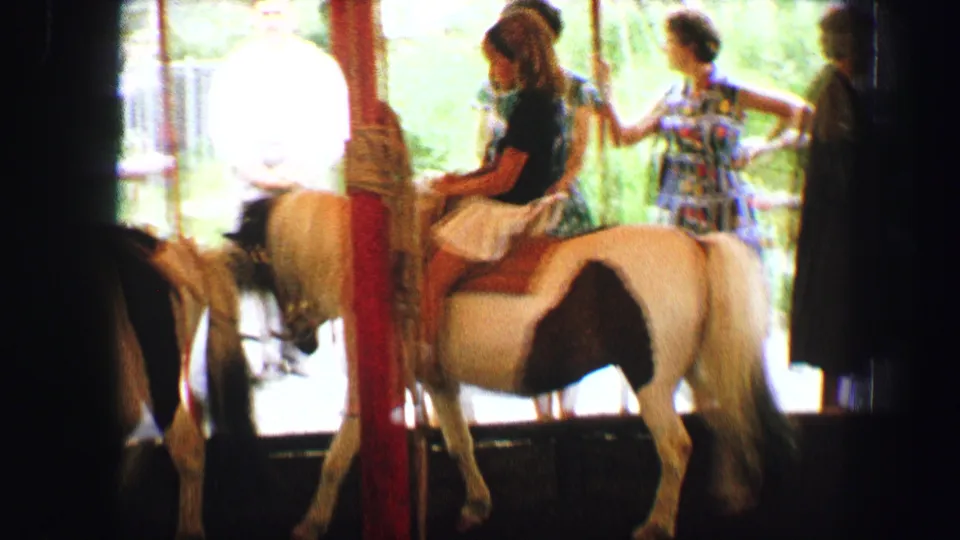 1963 Austria home-movie frame showing children near white ponies on a shaded outdoor pony ride with adults nearby.