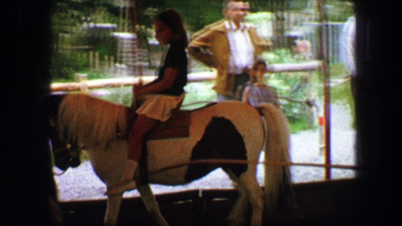 A child rides a white pony while adults and another child stand near a railing.