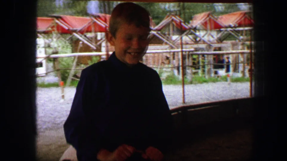 A boy stands in the shaded foreground of the 1963 Austrian pony ride scene, with railings and red canopy structures behind him.