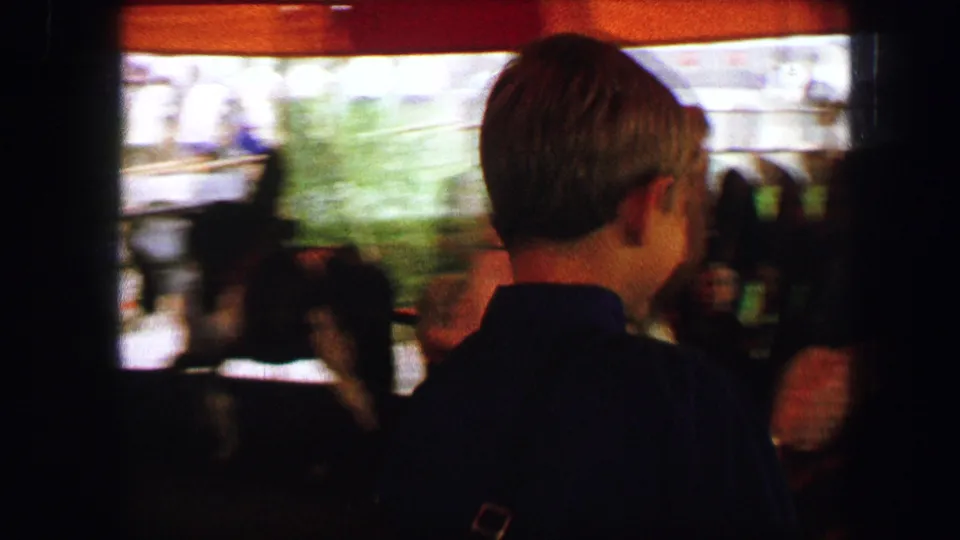 A boy seen from behind stands close to the moving pony ride, with blurred riders, ponies, and warm 8mm color in the frame.