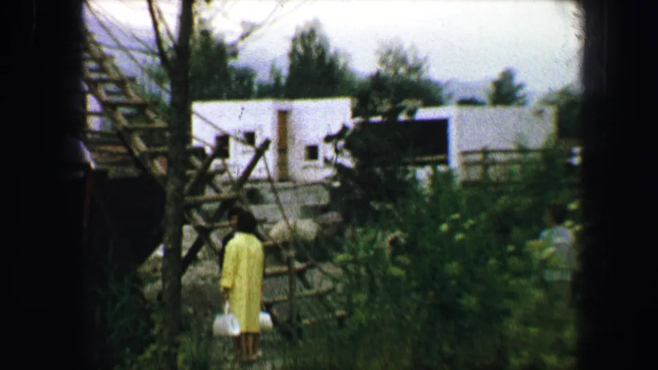A woman in a yellow coat stands near a rustic wooden play structure in a green Austrian garden setting, 1963.