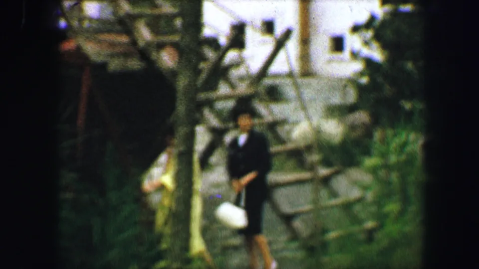 Two women, one in a light yellow coat and one in dark clothing, pass a wooden playground structure in 1963 Austria.