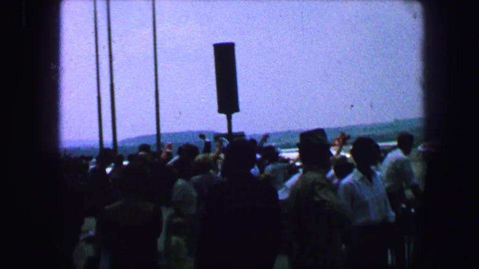 Austria 1963 airfield crowd seen in silhouette near vertical posts, with runway area and distant hills beyond.