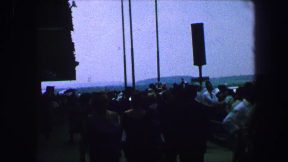 Austria 1963 home-movie view of spectators clustered by posts at an airfield, shot from a shaded or underexposed position.