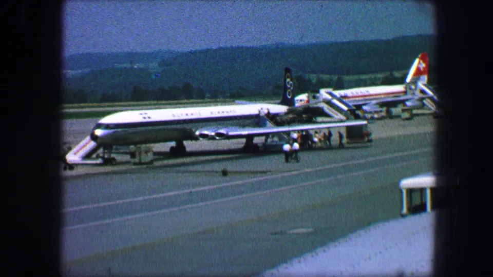 Austria 1963 frame showing a large passenger plane on the tarmac, with stairs, small figures, and wooded hills in the distance.