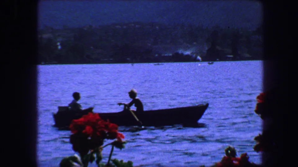 1963 Lucerne waterfront view showing a rowboat on blue water, red flowers near the window, and a hazy shoreline beyond.
