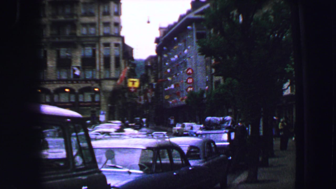 1963 8mm view of a Lucerne street with classic cars, pedestrians, buildings, and an overcast urban background.