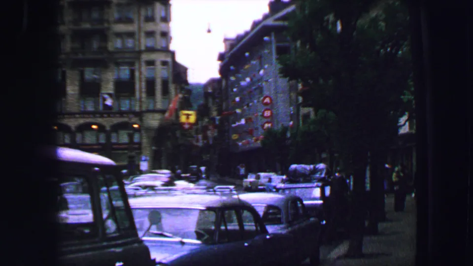1963 8mm view of a Lucerne street with classic cars, pedestrians, buildings, and an overcast urban background.