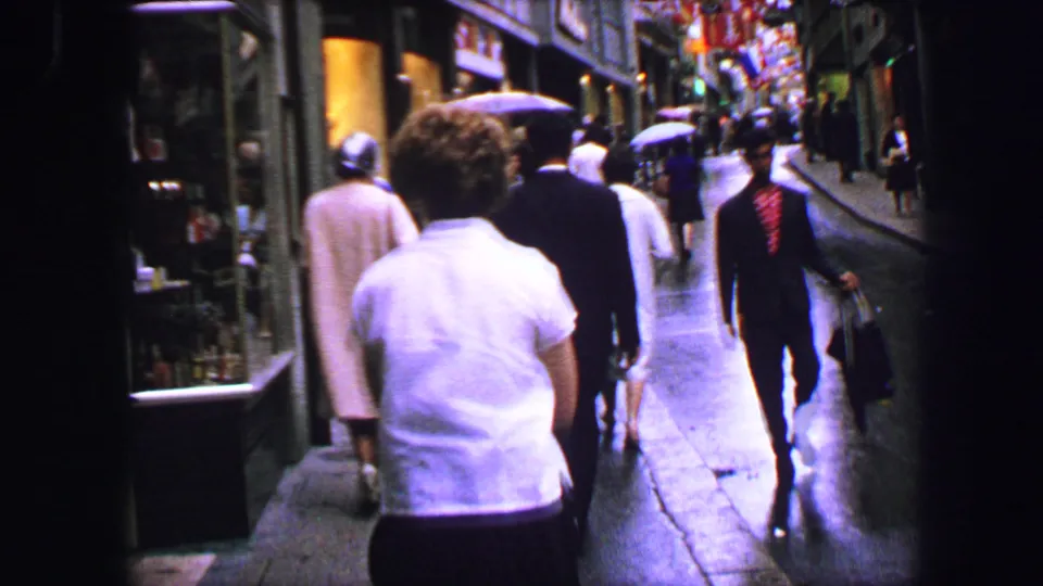 1963 Lucerne sidewalk view with pedestrians walking uphill beside shop windows, flags, and wet pavement.
