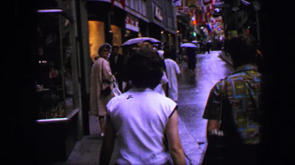 Lucerne street scene from 1963 showing pedestrians from behind on a narrow sidewalk with shopfronts and flags ahead.