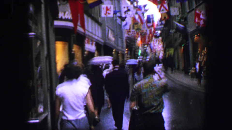 1963 8mm frame of people walking uphill on a wet Lucerne street lined with shopfronts and colorful flags.