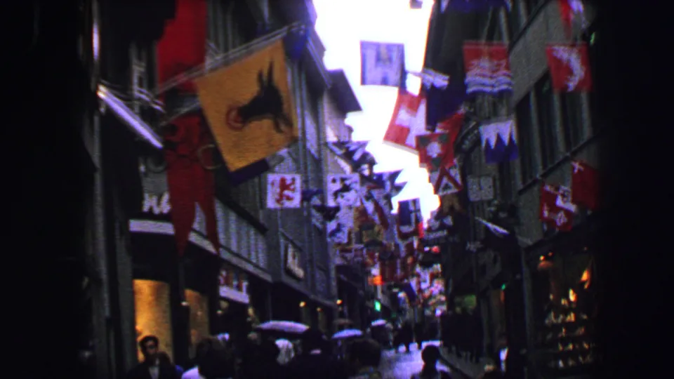1963 Lucerne street scene with overhead flags, dark shopfronts, pedestrians, umbrellas, and wet pavement.