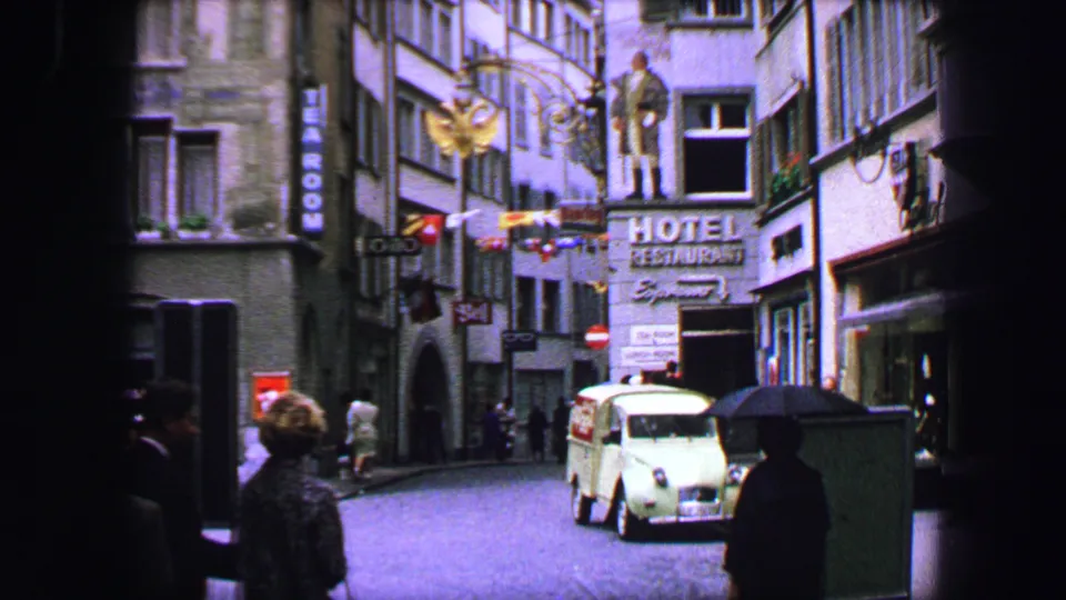 Lucerne street view from 1963 showing hotel signage, pedestrians with umbrellas, a light-colored period car, and historic facades.