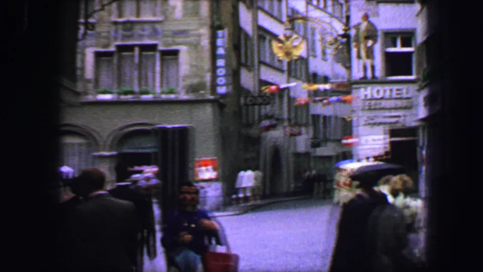 1963 Lucerne street frame with hotel signage, hanging flags, pedestrians, umbrellas, and a dim handheld 8mm view.