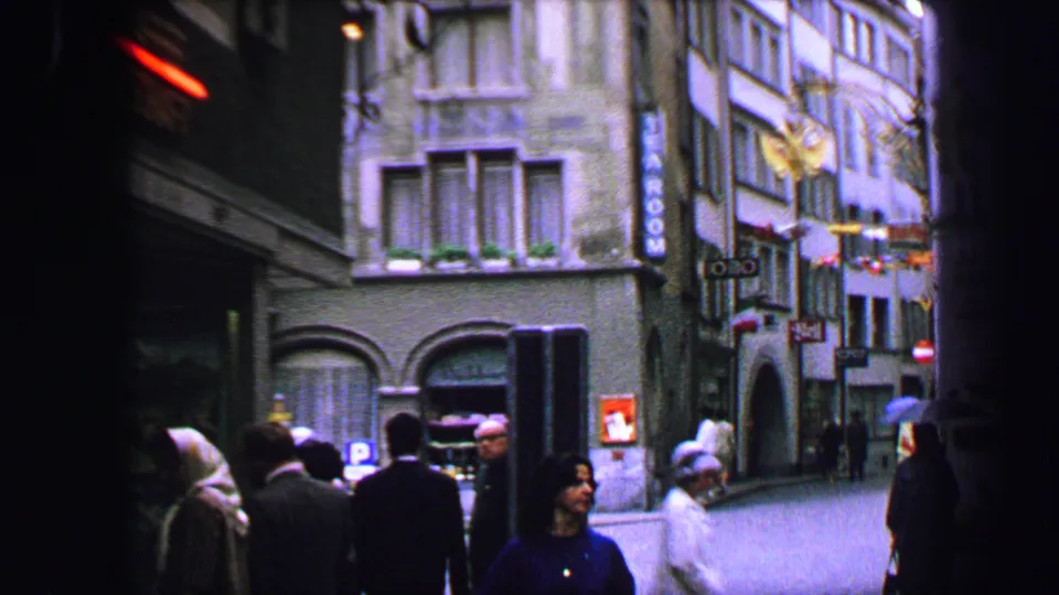 1963 Lucerne street view with pedestrians, old-town facades, hotel signage, and a red neon corner light in 8mm color footage.