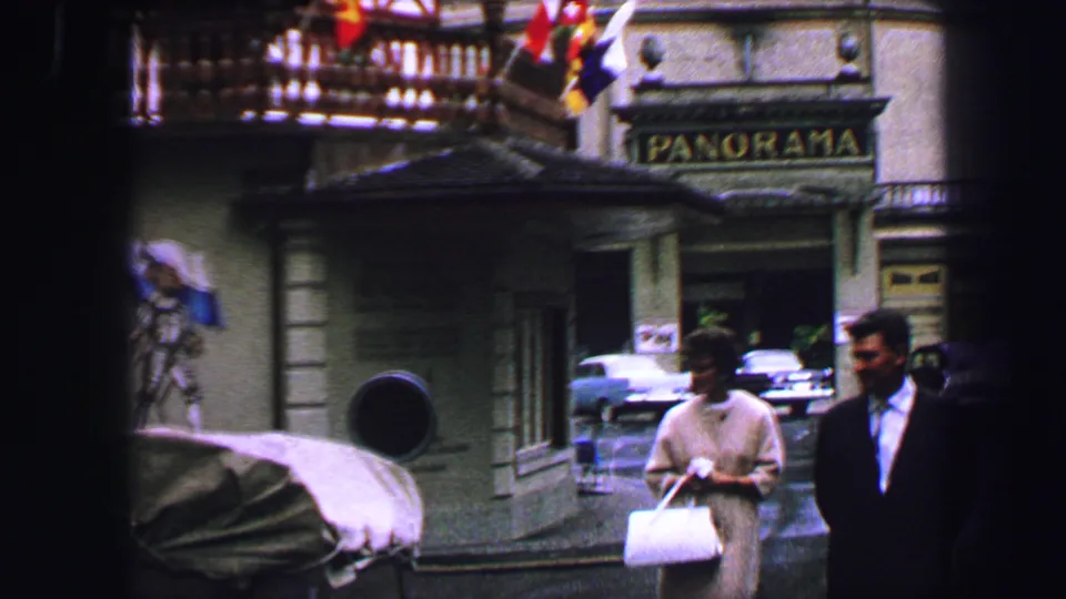 Lucerne street corner in 1963 with pedestrians, a Panorama sign, parked cars, stone facade, and flags near a building entrance.