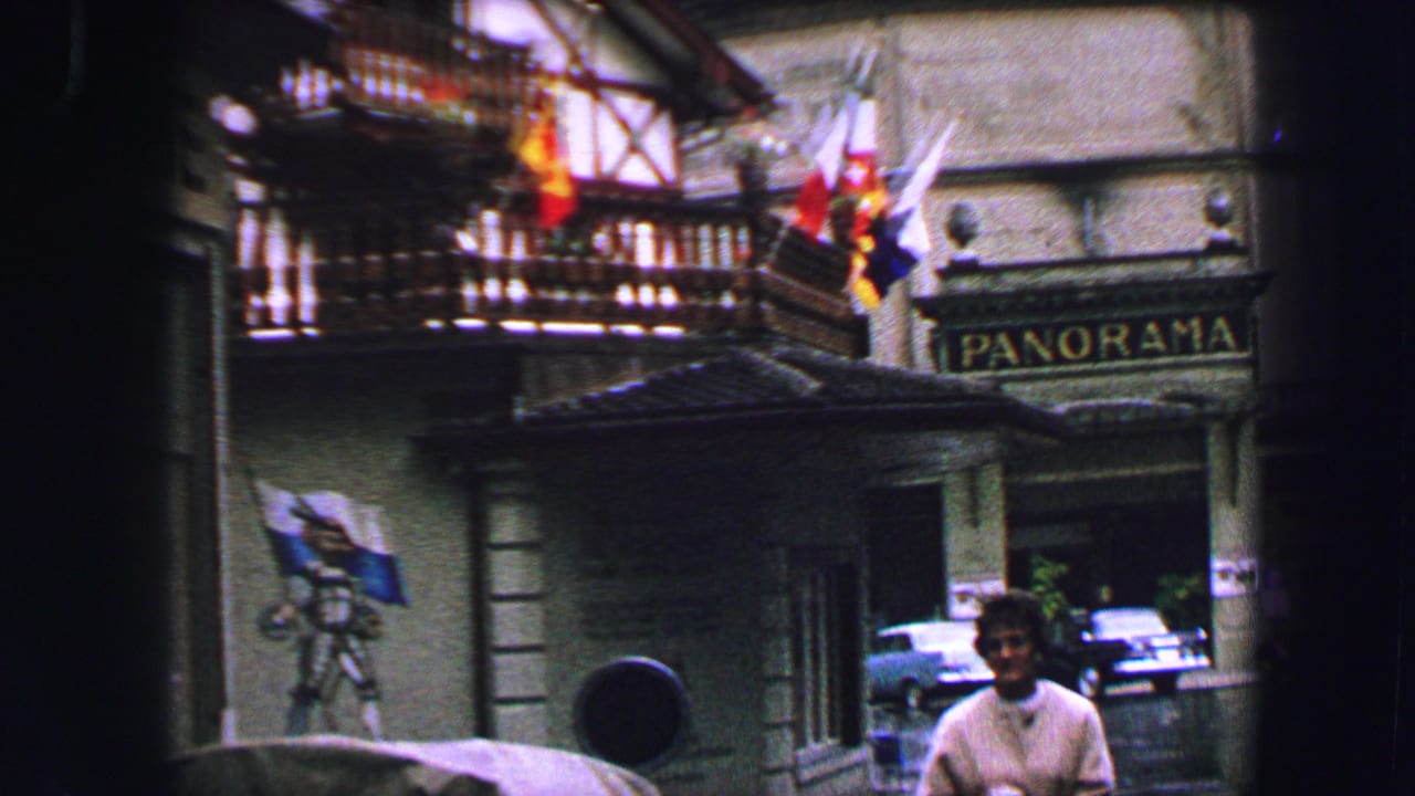 1963 Lucerne street view with a man below a traditional building, hanging flags, parked cars, and a PANORAMA sign.