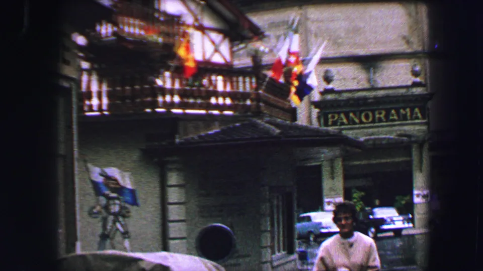 1963 Lucerne street view with a man below a traditional building, hanging flags, parked cars, and a PANORAMA sign.