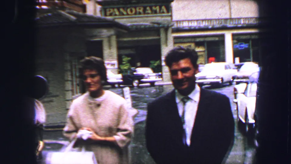 Two adults face the camera on a Lucerne street, with parked cars and a PANORAMA storefront sign behind them.