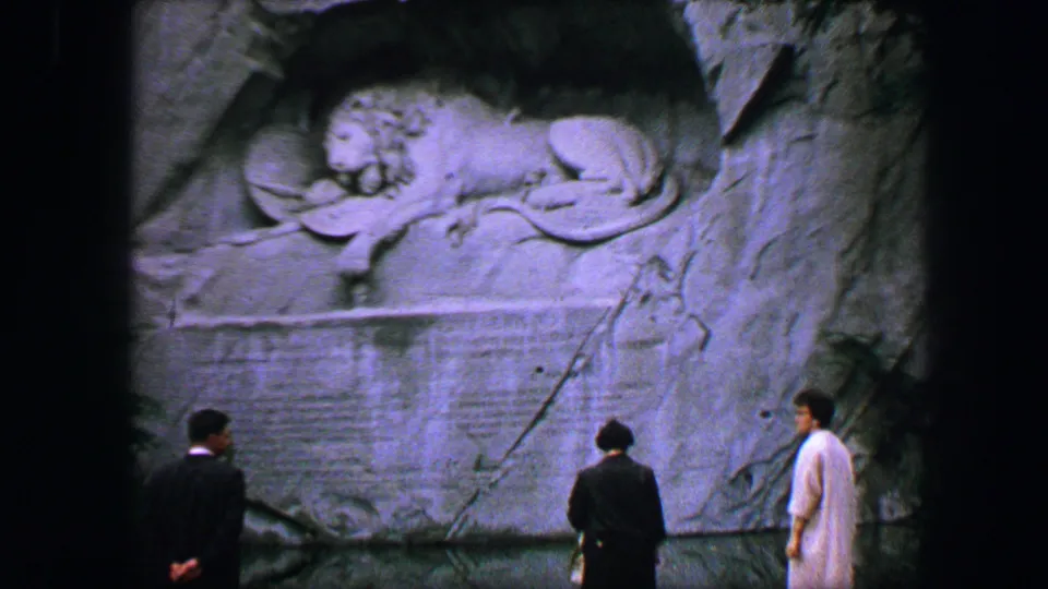 1963 Lucerne home-movie frame showing three visitors viewing the carved Lion Monument memorial in the rock wall.