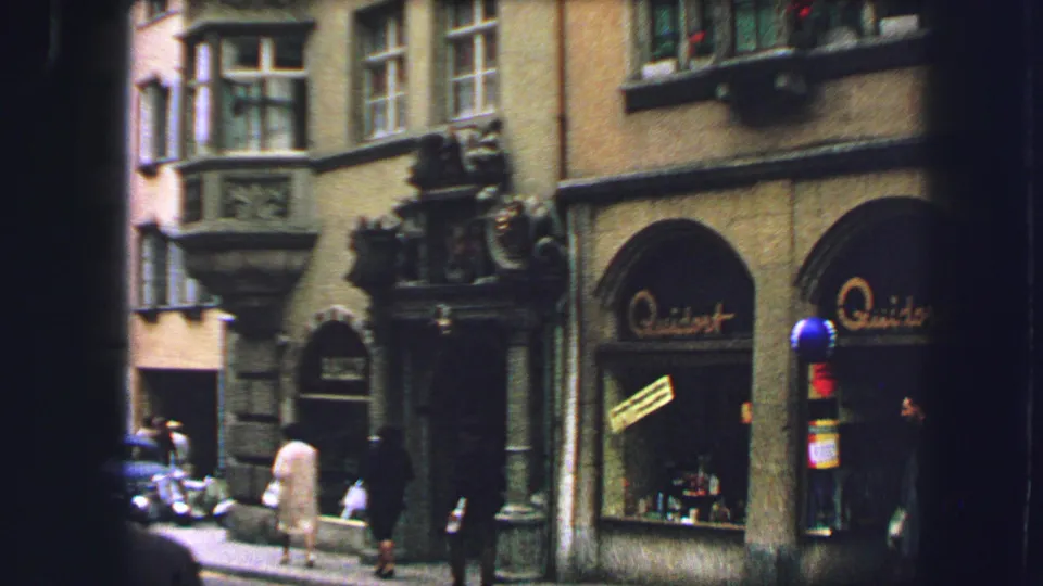 1963 Lucerne street view with pedestrians near an ornate facade, arched storefronts, and a small parked car.
