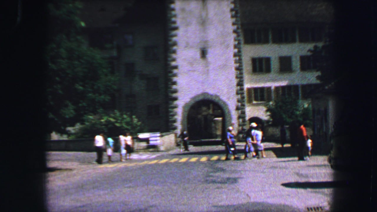 1963 Lucerne street view with pedestrians gathered near a yellow-striped crosswalk beside a stone arched gateway and shaded buildings.