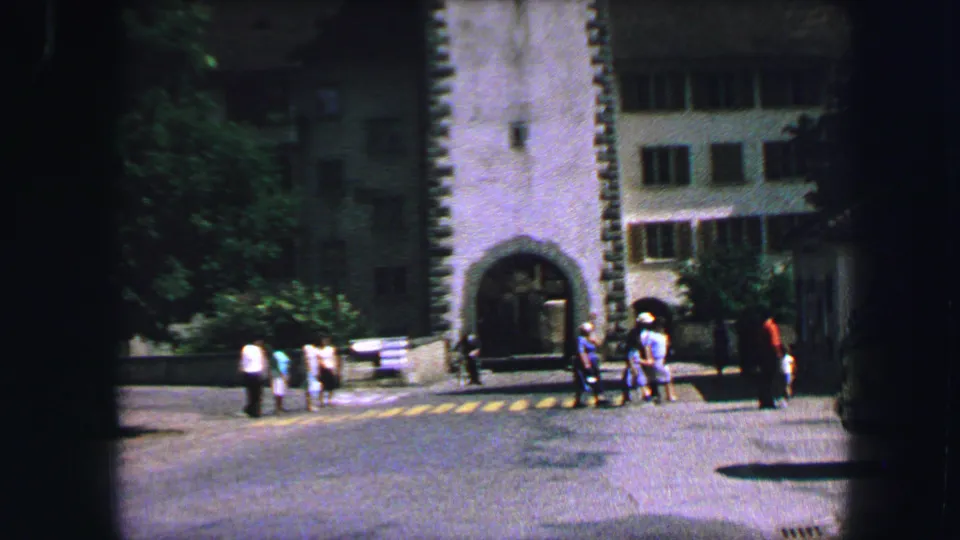 1963 Lucerne street view with pedestrians gathered near a yellow-striped crosswalk beside a stone arched gateway and shaded buildings.