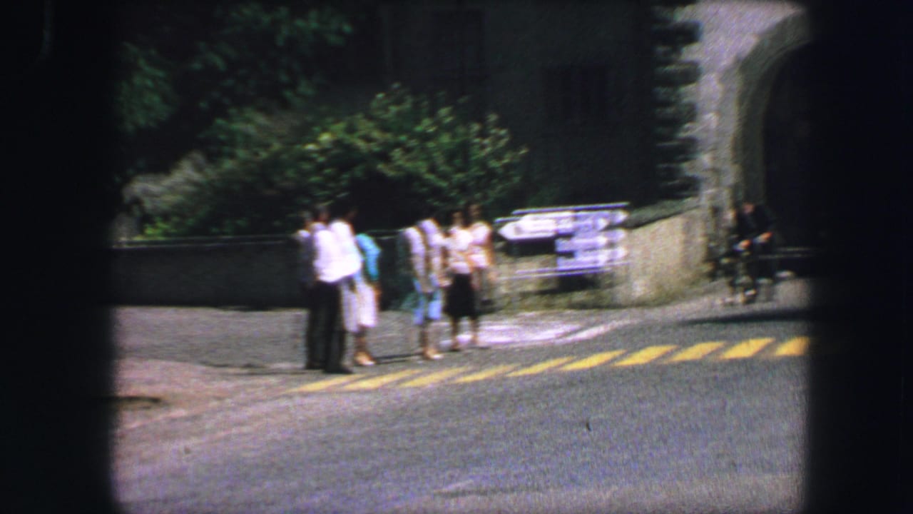 People wait beside a crosswalk near directional road signs and a curved stone wall.