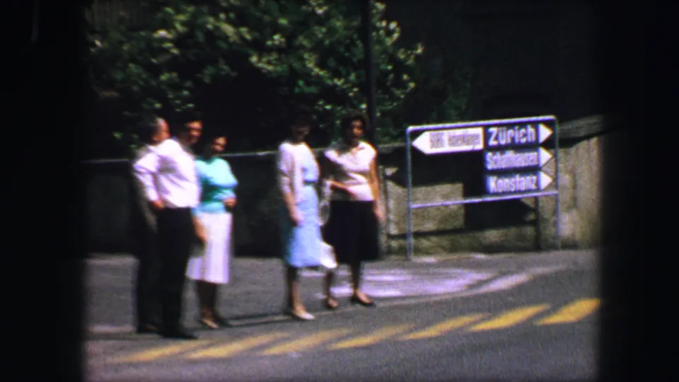 Three pedestrians stand by a Lucerne crosswalk near readable directional signs including Zurich, Sarnen, and Kussnacht.